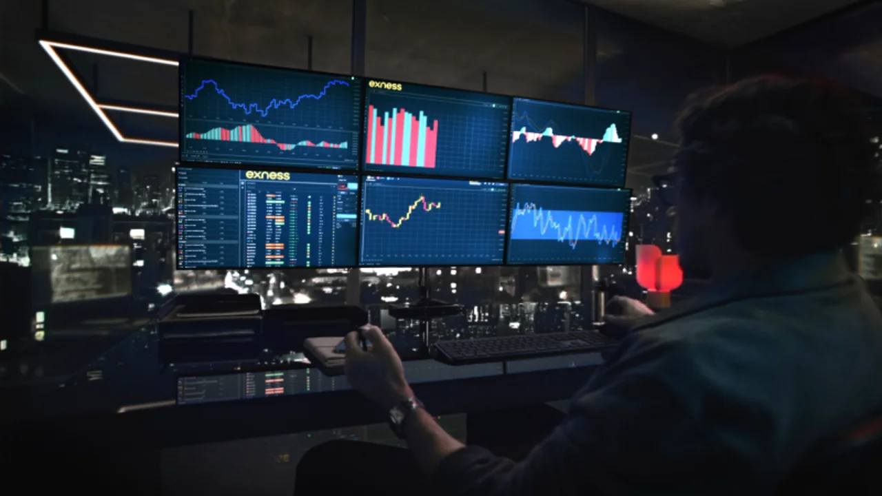 Trader operando con múltiples pantallas en un entorno nocturno, gráficos y plataformas de trading activas