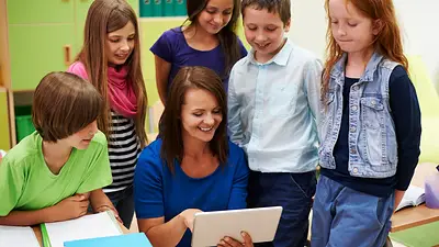 Docente usando una tablet junto a un grupo de alumnos en un aula Docente usando una tablet junto a un grupo de alumnos en un aula